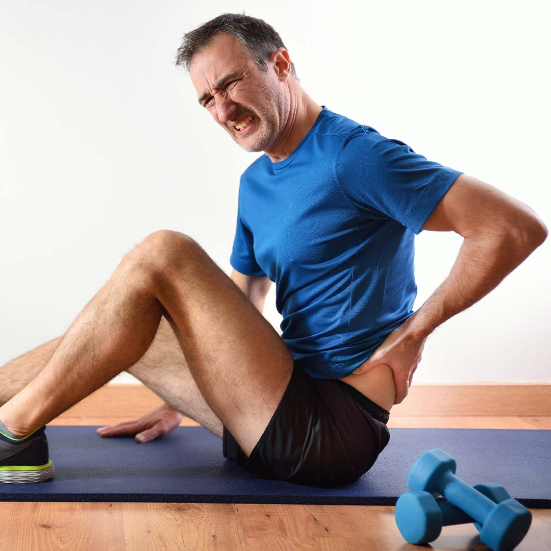 Homem com dor lombar após treino, sentado no tapete com halteres ao lado. Destaque para prevenção e fortalecimento.